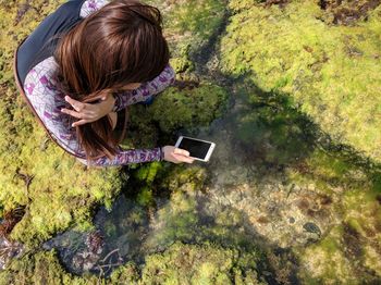 High angle view of young woman photographing lake while crouching on field