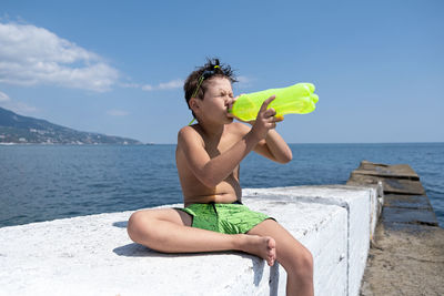 Boy drinking water sitting against sea