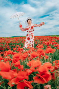 Happy woman in summer dress with a umbrella dances in the field with blooming poppies against a sky