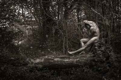Side view of naked man sitting on fallen tree in forest