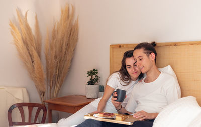 Cute cuddling and snuggling young couple sitting in bed having healthy breakfast 