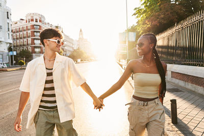 Happy multiracial man and woman in stylish clothes and sunglasses holding hands and looking at each other with smile while walking on sidewalk near fence and road at sundown in city