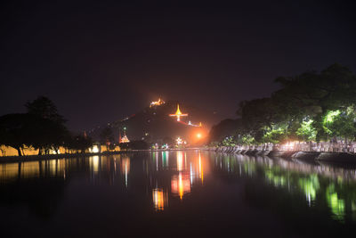 Illuminated buildings by lake against sky at night