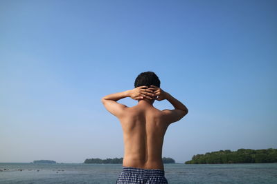 Rear view of shirtless man with hands behind head standing at beach against clear sky