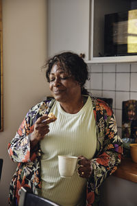 Mature woman with eyes closed having cookie and coffee in kitchen at home