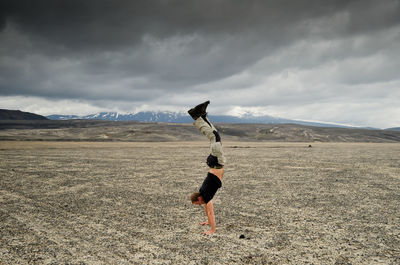 Full length of man practicing handstand on land against cloudy sky