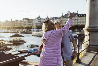 Smiling senior couple taking selfie while standing on bridge against lake in city