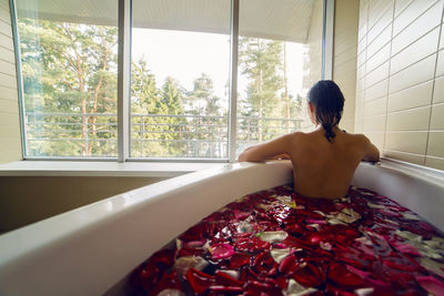 Young brunette woman with long hair white jacuzzi bath with rose petals, looking out the big window 