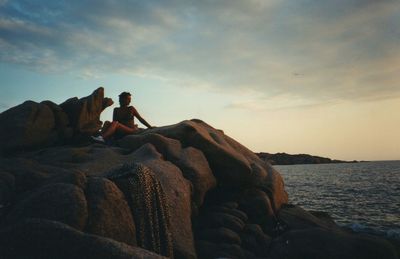 Man sitting on rock by sea against sky during sunset