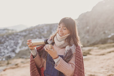 Woman pouring coffee from flask in cup while standing on land against mountain and sky