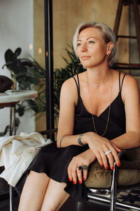 Blond stylish woman in black dress sitting in arm chair looking away