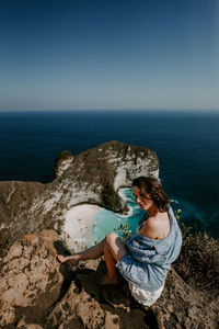 Rear view of woman sitting on rock by sea against sky