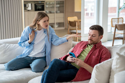 Offended wife reproaching uncaring husband in doing nothing, sitting, playing smarthphone games