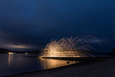 Light trails by river against sky at night