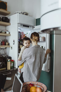 Rear view of mother carrying baby boy while opening refrigerator in kitchen