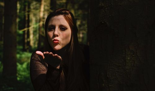 Portrait of young woman blowing kiss while standing behind tree trunk in forest