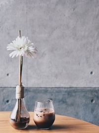 Close-up of white flower in vase with coffee on wooden table against wall