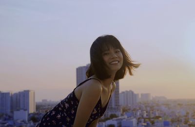 Portrait of smiling young woman against buildings in city at sunset