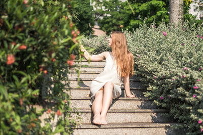 Beautiful young  woman in white dress sitting on the stairs in the park and looking at flowers. 