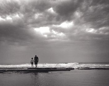 Mid distant view of boy with father walking at beach against cloudy sky