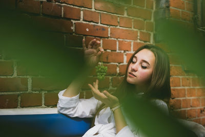 Young woman holding grapes while sitting on sofa against brick wall