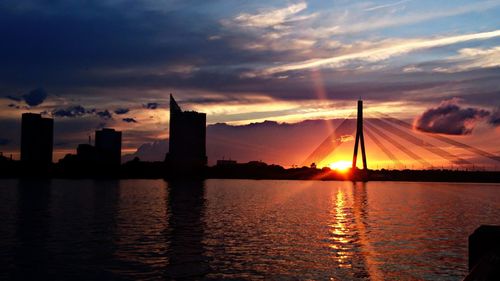 Silhouette buildings by river against sky during sunset