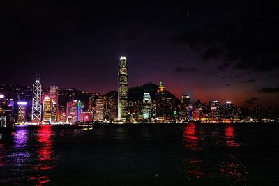 Illuminated buildings by river against sky at night