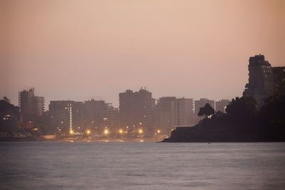 Silhouette buildings by sea against sky at sunset