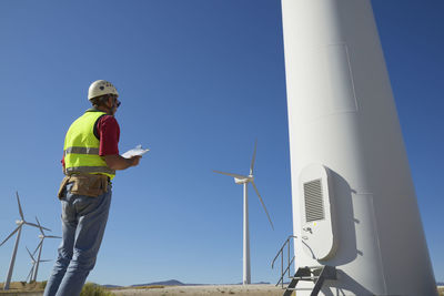 Low angle view of engineer holding clipboard while standing by windmills against sky