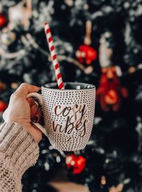 Close-up of woman hand holding drink in cup with text against christmas tree