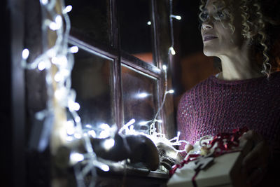 Smiling woman standing by illuminated window