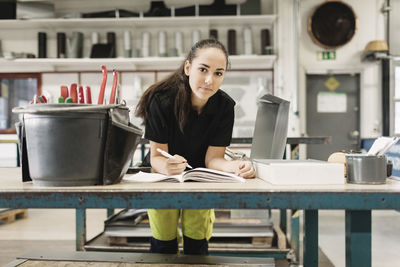 Portrait of confident young woman writing in book at workshop