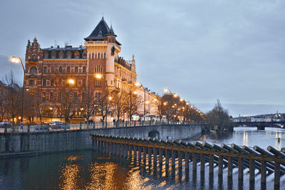 Illuminated buildings by river against sky in city