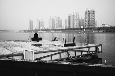 Rear view of man sitting by sea against buildings in city