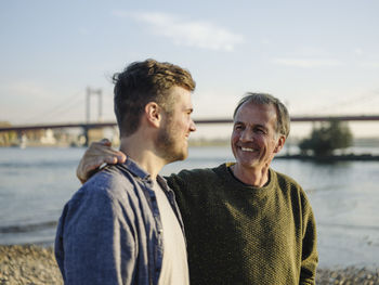 Happy father with arm around son at riverbank on sunny day