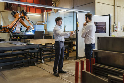 Young and mature businessman having discussion while standing against robotic machine at factory