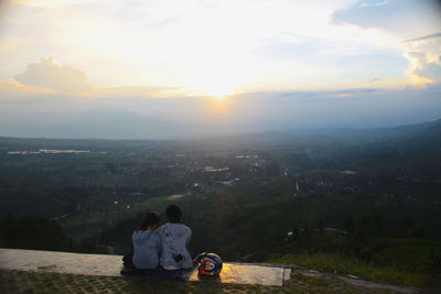 Rear view of boy sitting on mountain against sky during sunset