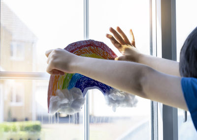 Cropped hands of boy holding rainbow drawing on window