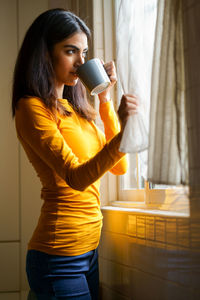 Young woman drinking coffee standing by wall indoors