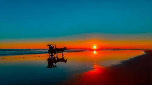 A silhouetted horse-drawn carriage at sunset time on the wet and reflecting sand beach in indonesia.