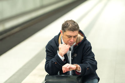 Man checking time while sitting with luggage at railroad station platform