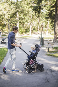 Full length of man walking while pushing son on baby carriage at public park