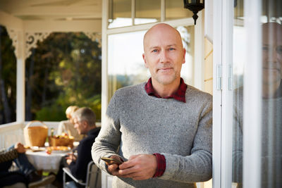 Portrait of confident bald mature man standing on porch with friends in background