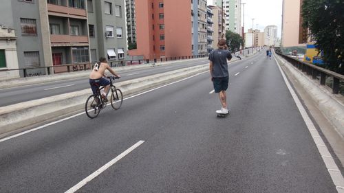 Shirtless man cycling with friend skateboarding at bridge in city