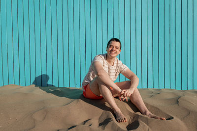 Young man sitting in sand beach smiling at camera in a turquoise wall