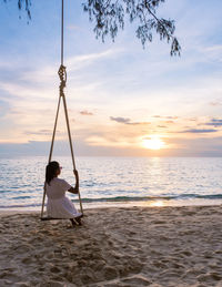 Rear view of woman swinging at beach against sky during sunset