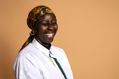 Cheerful african american female model in trendy headscarf and white shirt laughing with closed eyes on beige background in studio