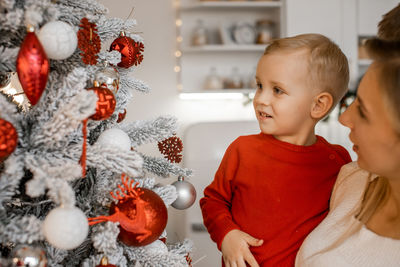 Charming mother holding her little son next to beautifully decorated christmas tree in the house