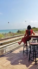 Rear view of woman sitting on retaining wall against sea