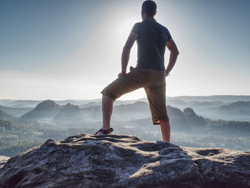 Sports man in outoor clothes and boots walking on rocky peak of mountain and look on nice fog view